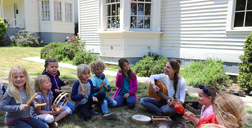 Unterricht draußen: eine Lehrerin und ihre Schülerinnen und Schüler sitzen auf einer Wiese und musizieren gemeinsam